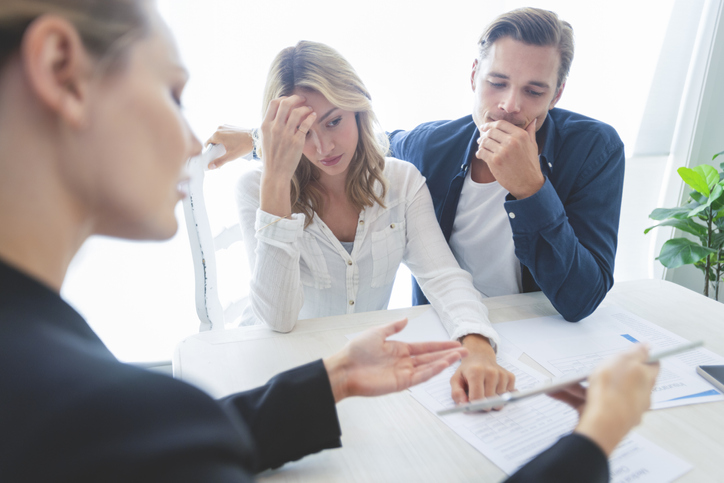 Real estate agent with couple looking through documents. The agent is holding a digital tablet showing it to the clients. Couple are casually dressed. They are looking concerned and worried and upset. Over the shoulder view.